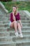 Blonde in light blue high-rise shorts, red plaid shirt, sneakers sitting on the stairs outside, looks to the left. vertical