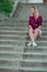 Blonde in light blue high-rise shorts, red plaid shirt, sneakers sitting on the stairs outside, looks to the left. vertical