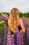 A blonde with long wavy hair stands with her back to the camera in lupine fields.