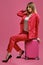 Blonde model in red pantsuit, white blouse, high black heels. She is sitting on her suitcase against pink background