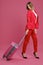 Blonde model in red pantsuit, white blouse, high black heels. She smiling, posing with suitcase on pink background