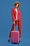 Blonde model in straw hat, white blouse, red pantsuit, high black heels. She is leaning on a handle of pink suitcase