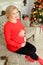 Blonde pregnant woman wearing red sweater hugging bely and sitting on sofa near Christmas tree.