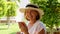 blonde senora woman, 50-55 years old, in a hat and a white dress, talks using a smartphone and holds a cup of coffee in