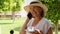 blonde senora woman, 50-55 years old, in a hat and a white dress, talks using a smartphone and holds a cup of coffee in