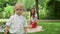 Blonde toddler covering face with hands. Family sitting on blanket in park.