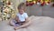 Blonde toddler girl lying on a bed in christmas decorated room and play with her digital tablet.