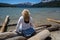 Blonde woman with back facing camera looks out to the Sawtooth Mountains while sitting on logs at Redfish Lake