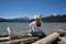 Blonde woman with back facing camera looks out to the Sawtooth Mountains while sitting on logs at Redfish Lake. Wind blows her