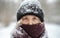 Blonde woman in beanie hat and nose covered in scarf looks at camera in winter snowfall. Nordic winter seasonal portrait, tranquil