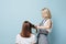 Blonde woman hairdresser straightening client`s hair with flat iron over blue