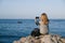 Blonde woman on her back sitting on a rock by the sea makes a photograph with the mobile phone of a sailboat sailing a sunny day