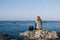 A blonde woman on her back sitting on a rock by the sea watching a sailboat sail a sunny day