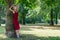 Blonde woman hugging a tree in park. Young girl in a red dress resting in nature, leaned against a tree.