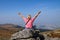 A blonde woman in a pink t-shirt sits on the top of a mountain raising her hands up