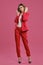 Blonde woman in red pantsuit, white blouse, high black heels. Posing standing against pink studio background. Beauty