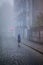 A blonde woman stands on the sidewalk at a crossroads in the dense autumn morning fog.
