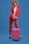 Blonde woman in straw hat, white blouse, red pantsuit, high black heels. She is leaning on a handle of pink suitcase