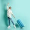 Blonde woman in white sweater, jeans and sneakers. She raised her hand up, smiling, holding suitcase, posing on blue background