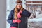 Blonde woman in winter hat and goggles holding a snowboard while sitting on a railing near the snowy sea coast