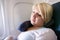 A blonde young adult woman uses a neck pillow to sleep and nap on a long airplane flight. Woman is in the window seat