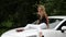 Blonde young woman sitting on the hood of the white car in summer day. slow motion