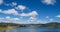 Blue calm deep water lake with gently sloping banks surrounded by hills with trees under a blue sky with small soft white clouds