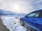 A blue car parked on the side of unpaved mountain road in the winter