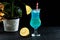 A blue cocktail glass decorated with Sicilian lemon, vase with a small palm tree next to the drink,