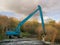 Blue color excavator working on deepening and cleaning a small river, Sunny day, Cloudy sky