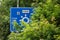 Blue direction traffic sign which cannot be read as it is overgrown by bush and trees. Neglected care about roads