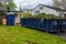 Blue dumpster filled with tree evergreen branches and a blue outhouse in the driveway of a house