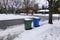 A blue and a green trash can covered with snow by the side of a snow covered, freshly plowed street waiting for garbage pickup