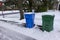 A blue and a green trash can covered with snow by the side of a snow covered, freshly plowed street waiting for garbage pickup