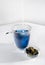 Blue herbal Butterfly pea flower tea in a glass mug on white background. A female hand pours milk from into tea. Next