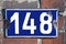 A blue house number plaque fixed on a brick wall, showing the number one hundred forty eight