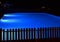 Blue illuminated pool at a night with a wooden black fence in foreground.