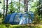 Blue large four-seater camping tent stands in shade of pine forest, weather is sunny. Summer camp, rest, hike.