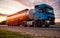 A blue modern truck with a semi-trailer tanker transports dangerous goods against the backdrop of the evening sunset. The concept