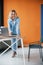 In blue official clothes. Businesswoman with curly blonde hair indoors in room with orange colored wall and wooden table