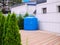 A blue plastic water tank stands in a backyard surrounded by thujas