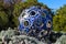 A blue and silver spherical aluminum sculpture in the garden surrounded by lush green plants and trees