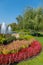 A blue sky above a fountain in a pond and a flower bed with a variety of colors