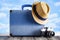 Blue suitcase, summer hat and photo camera on wooden table and beach sand background