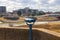 A blue telescope in front of a brown concrete wall near Wolf Creek Harbor surrounded by yellow winter grass with office buildings