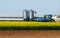 Blue tractor in the middle of a yellow canola field, with an agricultural building and two metal silos on behind.