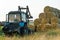 The blue tractor uses a hydraulic manipulator to stack round bales of hay in pyramids for storage and drying. Machinery works in