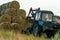 The blue tractor uses a hydraulic manipulator to stack round bales of hay in pyramids for storage and drying. Machinery works in