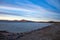 Blue water filled lake dam with rocky foreshore and small hills in background against blue sky