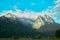 Blue-white sky above the Zugspitze with wooded mountain slopes and a meadow with barns and sheds